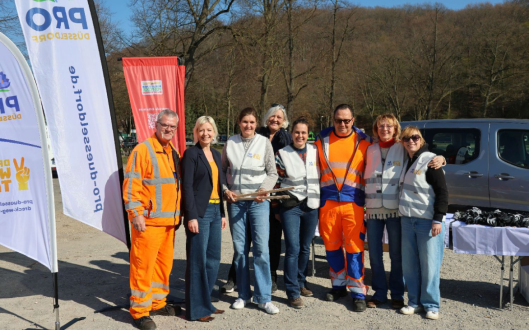Initiatoren und Unterstützer:innen des Dreck-weg-Tags 2026 vor einem Ausgabestand mit Handschuhen, Westen und Zangen, im Hintergrund Fahnen mit dem Pro Düsseldorf Logo, Dachorganisation des Dreck-weg-Tages