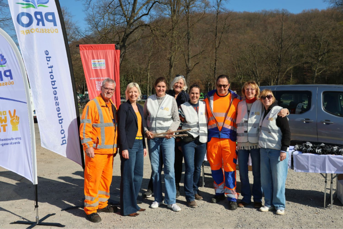 Initiatoren und Unterstützer:innen des Dreck-weg-Tags 2026 vor einem Ausgabestand mit Handschuhen, Westen und Zangen, im Hintergrund Fahnen mit dem Pro Düsseldorf Logo, Dachorganisation des Dreck-weg-Tages